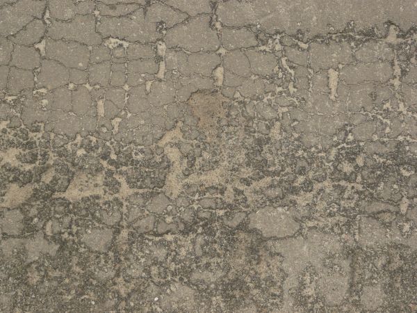 Grey asphalt texture covered in thick, shallow cracks and areas of dark, crumbling rock.